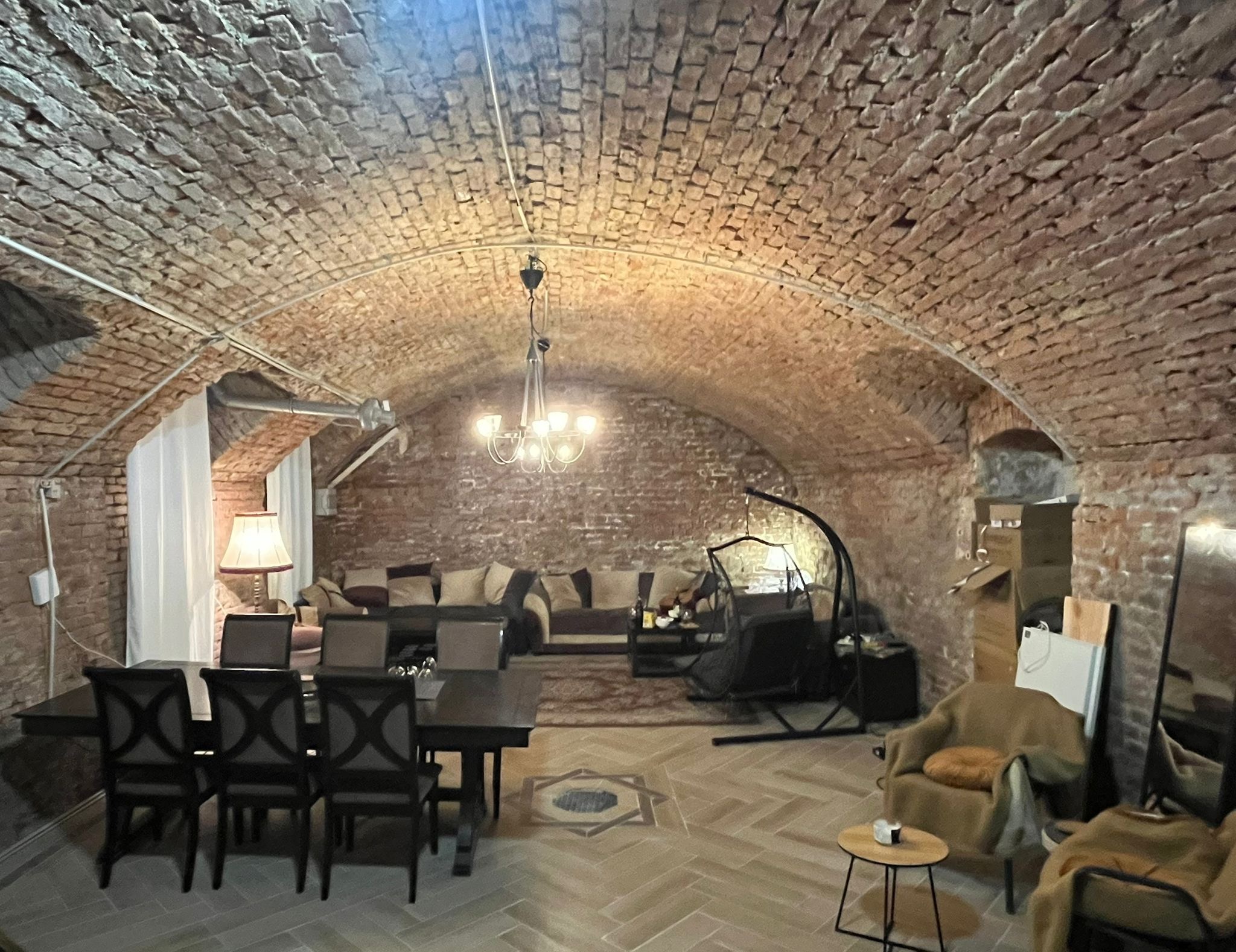 A brick barrel-vaulted cellar room lit by a chandelier, with a long dining table and lounge seating.